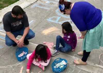 two adults watching two children coloring with chalk on sidewalk